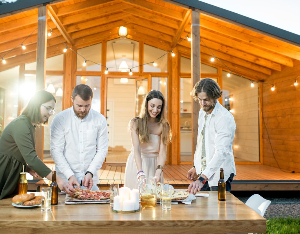 Friends having a dinner with pizza on the backyard of the modern house decorated with lights during the evening time outdoors